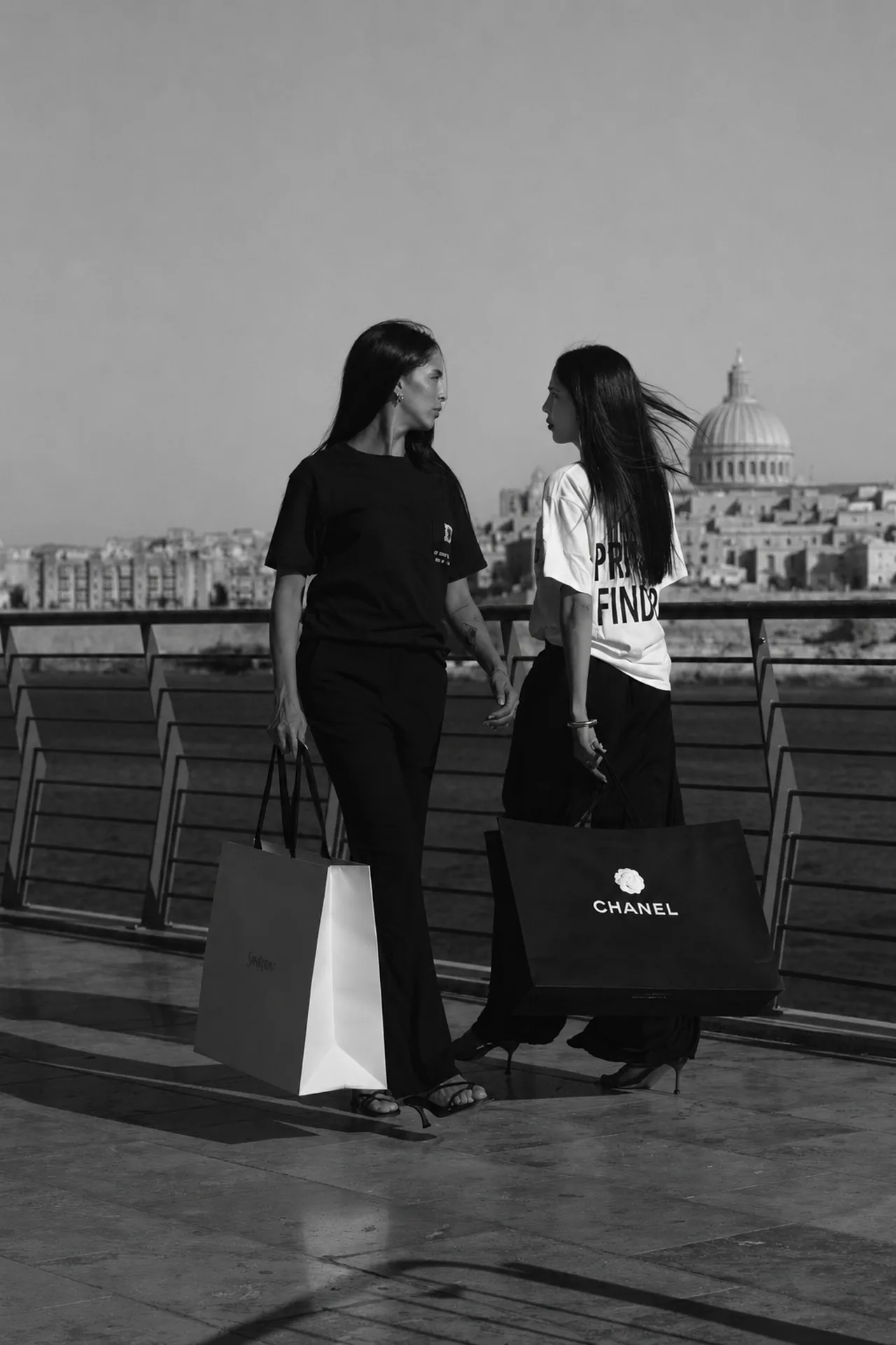 Two women in The Private Finder tees on the Valletta waterfront, luxury bags in hand.